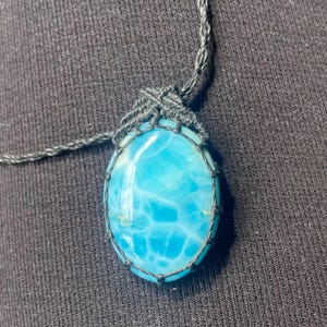 Puede incluir: Un collar con colgante de piedra preciosa ovalada azul. La piedra preciosa es de color azul claro con un patrón único, encerrada en un engaste de macramé negro. El cordón del collar también es negro.
