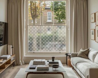May include: A living room with a large window featuring a white patterned window film. The room has a beige sofa, a coffee table with books and a vase, and a wooden entertainment center with a television.