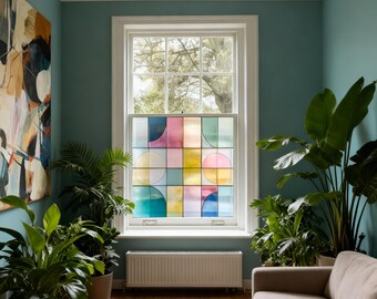 May include: A window with a colorful geometric design, featuring squares and semi-circles in shades of pink, blue, yellow, and green. The window is set in a white frame, with lush green plants and a beige sofa in the room.