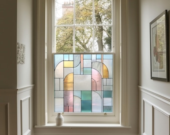 Op de afbeelding: Een raam met een gebrandschilderd glazen paneel met geometrische vormen in blauw, geel, roze en groen. Het raam heeft een witte lijst en een kleine witte vaas staat op de vensterbank.