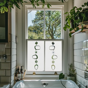Könnte beinhalten: Ein Badezimmerfenster mit weißem Rahmen und Milchglasscheiben. Die Paneele weisen ein sich wiederholendes Muster aus grünen und grauen abstrakten Formen auf. Das Fenster blickt auf einen grünen Baum. Das Badezimmer hat weiße U-Bahn-Fliesen und eine weiße Badewanne.