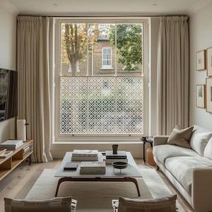 May include: A bright living room with a large window adorned with a patterned film. Beige curtains frame the window, and a white sofa and coffee table are in the foreground. The room features wooden flooring and neutral walls, creating a light and airy space.