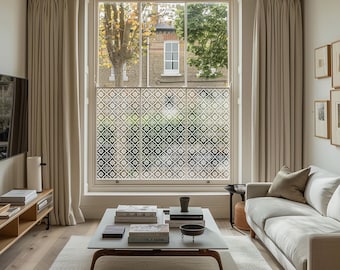 May include: A living room with a large window featuring a decorative patterned film. Beige curtains frame the window, and a white sofa and coffee table are in the foreground. The room has hardwood floors and neutral-toned walls, creating a bright and airy atmosphere.