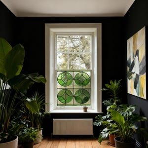 May include: Interior shot of a room with a large window. The window has a stained glass design with green circular patterns. The room features black walls, wooden flooring, and several potted plants. A painting hangs on the wall.