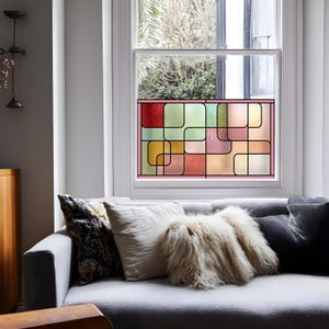 Könnte beinhalten: Ein Buntglasfenster mit einem geometrischen Muster aus Quadraten und Rechtecken in Rot, Gelb, Grün und Rosa. Das Fenster ist schwarz umrandet und in einem weißen Fensterrahmen eingesetzt. Das Fenster befindet sich über einem grauen Sofa mit Kissen.