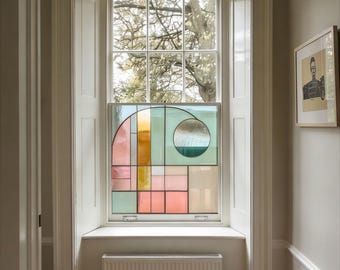 May include: A stained glass window featuring a geometric design in pastel shades of pink, green, and orange. The window is set within a white-framed window with shutters. The artwork is a decorative feature in a room.