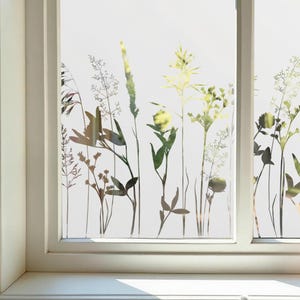 Puede incluir: Una ventana blanca con un diseño de vidrio esmerilado que presenta siluetas de varias plantas y hierbas en tonos verdes, marrones y amarillos. La ventana está enmarcada en blanco y se encuentra sobre una superficie de baldosas blancas.