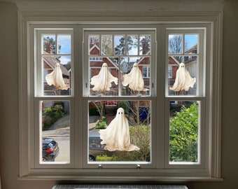 Puede incluir: Un marco de ventana blanco con seis decoraciones de fantasmas colgantes. Los fantasmas tienen un diseño simple con dos ojos de puntos negros y un cuerpo fluido similar a una sábana. La ventana da a una calle residencial con casas y árboles.