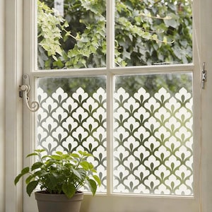 May include: A white-framed window with a decorative pattern on the glass. The pattern is a repeating design of white floral shapes. A potted plant sits on the windowsill. Green foliage is visible outside the window.