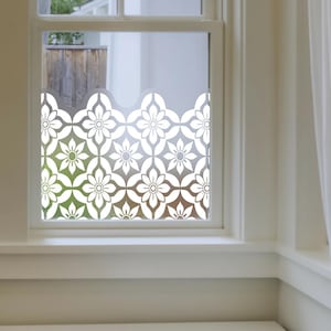 Könnte beinhalten: Weiße Fensterfolie mit Blumenmuster, die auf einem Fenster angebracht ist. Die Folie zeigt ein sich wiederholendes Muster aus stilisierten Blumen und Blättern. Das Fenster ist weiß umrahmt und bietet einen Blick auf einen Garten und einen weißen Vorhang.