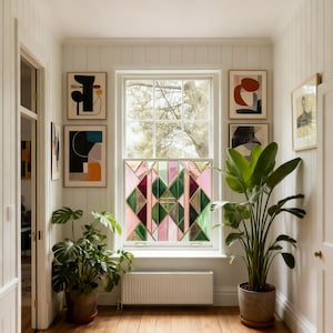 May include: A hallway with a window featuring a stained glass design in pink, green, and gold geometric shapes. The room has framed abstract art, potted plants, and a radiator. The walls are white with vertical paneling, and the floor is wood.