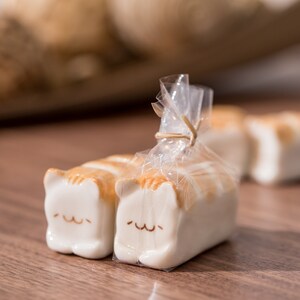 May include: Ceramic cat-shaped soap bars with white and orange stripes, packaged in clear cellophane. The cat soaps have a smiling face design. The background is blurred, with more cat soaps visible.
