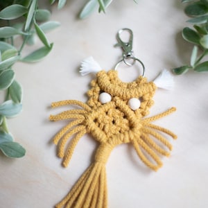 Peut inclure: Un porte-clés en macramé jaune avec un pompon blanc et des perles en bois pour les yeux. Le porte-clés est attaché à un anneau de clé argenté.