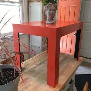 May include: A bright orange square side table with four thick legs and a smooth, solid surface. The table features a clean, block-like design with a matte finish. A grey stone-like bust planter containing green foliage sits on the table top.