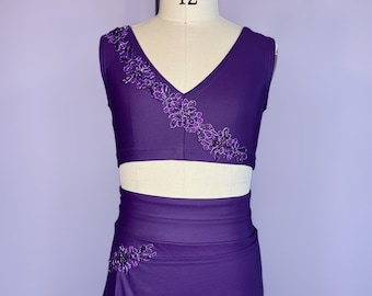 May include: A two-piece dance costume in a deep purple hue. The top is a V-neck crop top with floral embroidery. The skirt is a wrap-style design with matching floral detailing. The mannequin is wearing a size 12.