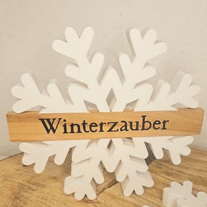 Può includere: Decorazione a forma di fiocco di neve in legno bianco con la scritta "Winterzauber" in lettere nere su una tavola di legno. Il fiocco di neve è appoggiato su una superficie di legno.