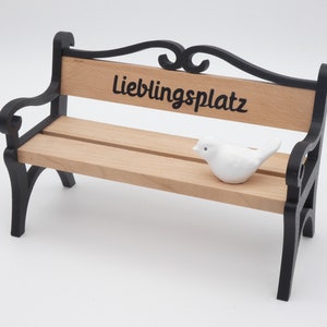Könnte beinhalten: Eine kleine Holzbank mit schwarzen Metallbeinen und einem dekorativen Schnörkeldesign auf der Rückenlehne. Die Bank hat ein Schild mit der Aufschrift "Lieblingsplatz" und ein weißer Keramikvogel sitzt darauf.