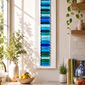 Puede incluir: Una pieza de arte mural vertical que presenta un panel rectangular blanco adornado con una columna única de piezas de vidrio apiladas, lisas y redondeadas, en varios tonos de azul, verde azulado y verde, creando un degradado de tonos fríos.