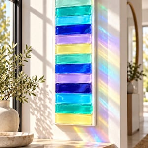 May include: A vertical rectangular wall art piece composed of stacked, translucent glass bars in shades of light green, turquoise, blue, purple, and yellow. Sunlight passing through the glass casts a vibrant rainbow light projection.