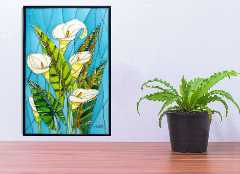 May include: A framed stained glass artwork featuring white calla lilies with green leaves on a blue background. The artwork is framed in black and has a signature in the bottom right corner.