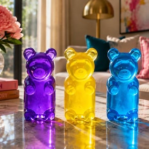 May include: Three colorful gummy bear-shaped decorative objects in purple, yellow, and blue. The translucent bears are displayed on a reflective surface, with a vase of pink flowers and books in the background. Home decor.