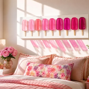 May include: A rectangular wall art piece featuring nine translucent popsicle sculptures arranged in a horizontal row. The popsicles transition in color from light pink to deep magenta, each mounted on a white rectangular board with wooden sticks.