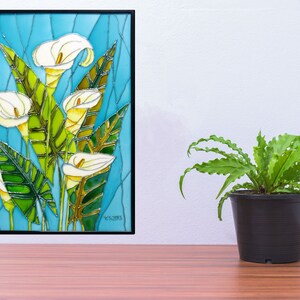 May include: A framed stained glass artwork featuring white calla lilies with green leaves on a blue background. The artwork is framed in black and has a signature in the bottom right corner.