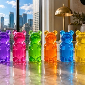 May include: Six colorful gummy bear-shaped containers, each approximately 4.5 inches tall, are arranged in a row. The colors include purple, pink, green, orange, blue, and yellow. The transparent containers sit on a reflective surface, with a cityscape visible in the background.