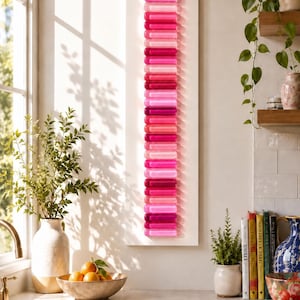 May include: A vertical wall art piece featuring a rectangular white panel adorned with a single column of numerous rounded, translucent glass-like capsules in varying shades of pink, ranging from light pastel to deep magenta, arranged in a repeating pattern.