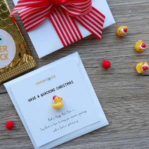 Könnte beinhalten: Eine weiße Karte mit dem Text "HAVE A QUACKING CHRISTMAS" und einer kleinen gelben Gummiente, die einen roten Weihnachtsmann-Hut trägt. Die Karte liegt auf einer Holzoberfläche mit einem roten Bommel und einer kleinen gelben Gummiente, die einen roten Weihnachtsmann-Hut trägt.
