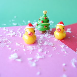 May include: Two yellow rubber duckies wearing red Santa hats, and a small green Christmas tree with red ornaments, on a pink and red surface with white confetti.