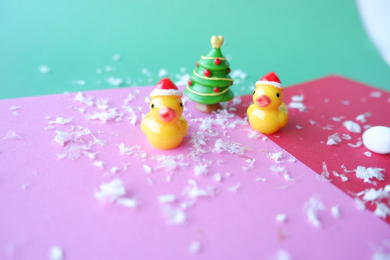 May include: Two yellow rubber duckies wearing red Santa hats, and a small green Christmas tree with red ornaments, on a pink and red surface with white confetti.