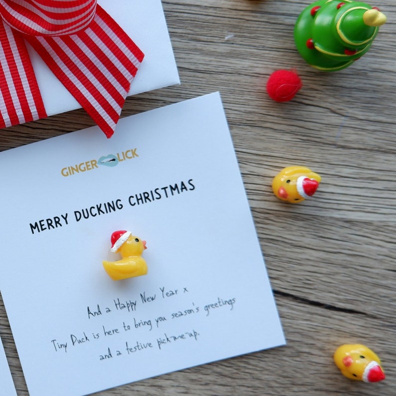 May include: A white card with the text "Merry Ducking Christmas" and "And a Happy New Year x Tiny Duck is here to bring you season's greetings and a festive pick me up."  The card has a small yellow rubber duck with a red Santa hat on it.