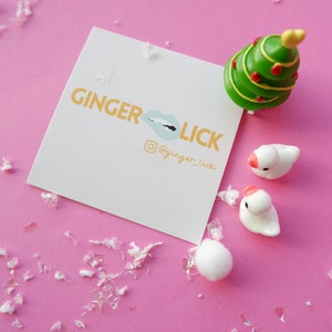 May include: A white card with the text "GINGER LICK" in orange letters and a blue lip graphic. The card also includes the text "@ginger_lick" in orange letters. The card is on a pink surface with white confetti and a small green Christmas tree and two white ceramic birds.