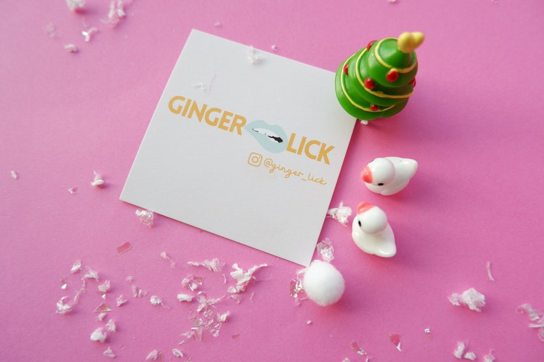 May include: A white card with the text "GINGER LICK" in orange letters and a blue lip graphic. The card also includes the text "@ginger_lick" in orange letters. The card is on a pink surface with white confetti and a small green Christmas tree and two white ceramic birds.