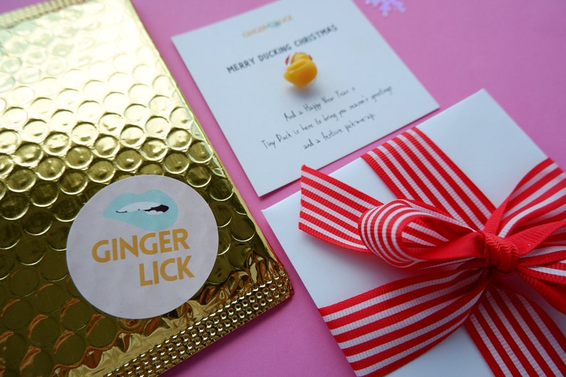 May include: A gold bubble mailer with a white sticker that says "GINGER LICK" with a blue and white lip design. A white card with red and white striped ribbon tied in a bow. The card says "MERRY DUCKING CHRISTMAS" and "And a Happy New Year. Tiny Duck is here to bring you mama's greeting and a festive pick-me-up."