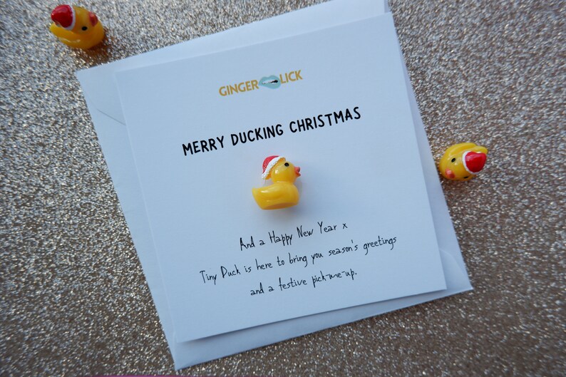 May include: A white card with the text "GINGERLICK" and "MERRY DUCKING CHRISTMAS" in black. A small yellow rubber duck wearing a red Santa hat is attached to the card. The text "And a Happy New Year x" is written in black ink below the duck. The text "Tiny Puck is here to bring you season's greetings and a festive pick me up." is written in black ink below the previous text.