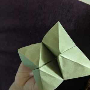 May include: A green origami fortune teller, also known as a cootie catcher, is folded into a pyramid shape.