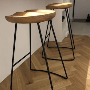 Puede incluir: Dos taburetes de bar de madera con patas de metal negro. Los asientos son de color marrón claro y tienen un diseño curvado. Las patas delgadas e inclinadas proporcionan una estética moderna. Los taburetes están sobre un suelo de madera.