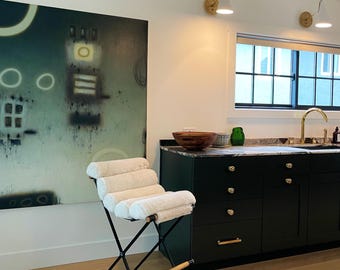 May include: A black and white abstract painting hangs on a white wall above a black and white folding stool with a white cushion. The stool is in front of a dark green kitchen island with gold hardware.