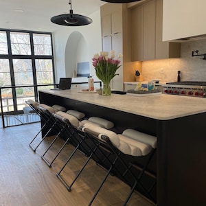 May include: A modern kitchen island with a black countertop and white bar stools. The island has a black base and a light gray countertop. The stools have white cushions and black metal frames.