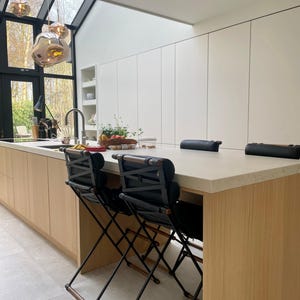 Pode incluir: Uma ilha de cozinha moderna com um balcão de madeira clara e dois banquetas de bar pretas com detalhes em cobre. A ilha está localizada em frente a uma grande janela com vista para as árvores.