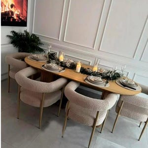 May include: A dining table with six beige upholstered chairs. The oval table is made of light wood and is set with plates, cutlery, and candles. The background includes a fireplace and decorative greenery.