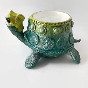 May include: A green and yellow ceramic turtle planter with a white interior. The turtle has a decorative design on its shell and a yellow flower on its head.
