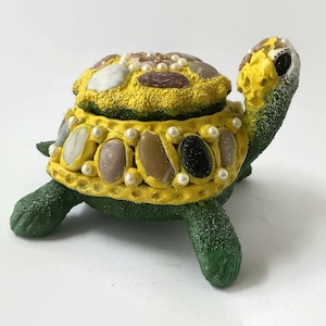 May include: A green and yellow turtle-shaped trinket box decorated with small stones and pearls. The box is open and the inside is lined with yellow.