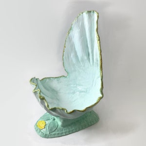 Könnte beinhalten: Eine hellblaue Keramikschale mit goldfarbenem Rand und grünem Sockel. Die Schale ist muschelförmig und hat eine kleine gelbe Blume, die auf den Sockel gemalt ist.