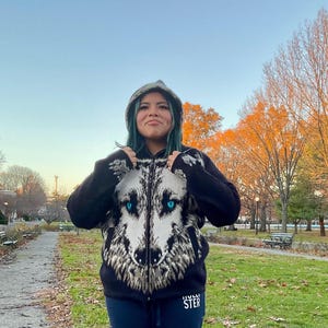 Op de afbeelding: Zwarte hoodie met ritssluiting met een gedetailleerd wolfsgezichtontwerp op de voorkant. De wolf heeft blauwe ogen en een wit en grijs vachtpatroon. De hoodie heeft een capuchon en het woord "HOLLI STER" staat onderaan.