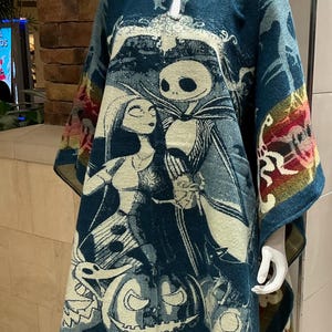 Pode incluir: Um poncho azul turquesa com capuz e borda de franja apresenta uma imagem impressa de Jack Skellington e Sally do filme O Estranho Mundo de Jack. A imagem também inclui abóboras e outras imagens de Halloween.