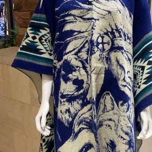 Może przedstawiać: Niebieskie poncho z motywem wilka. Poncho posiada kaptur z zapięciem na guzik i frędzle na dole. Projekt zawiera twarz wilka i geometryczne wzory w kolorze białym, turkusowym i czarnym.