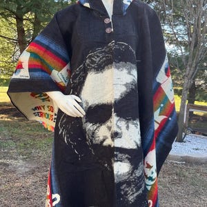Pode incluir: Poncho preto com um grande gráfico de um rosto. O poncho tem um capuz e dois botões. As bordas são adornadas com franjas coloridas. As palavras "WHY SO SERIOUS?" são impressas no poncho.